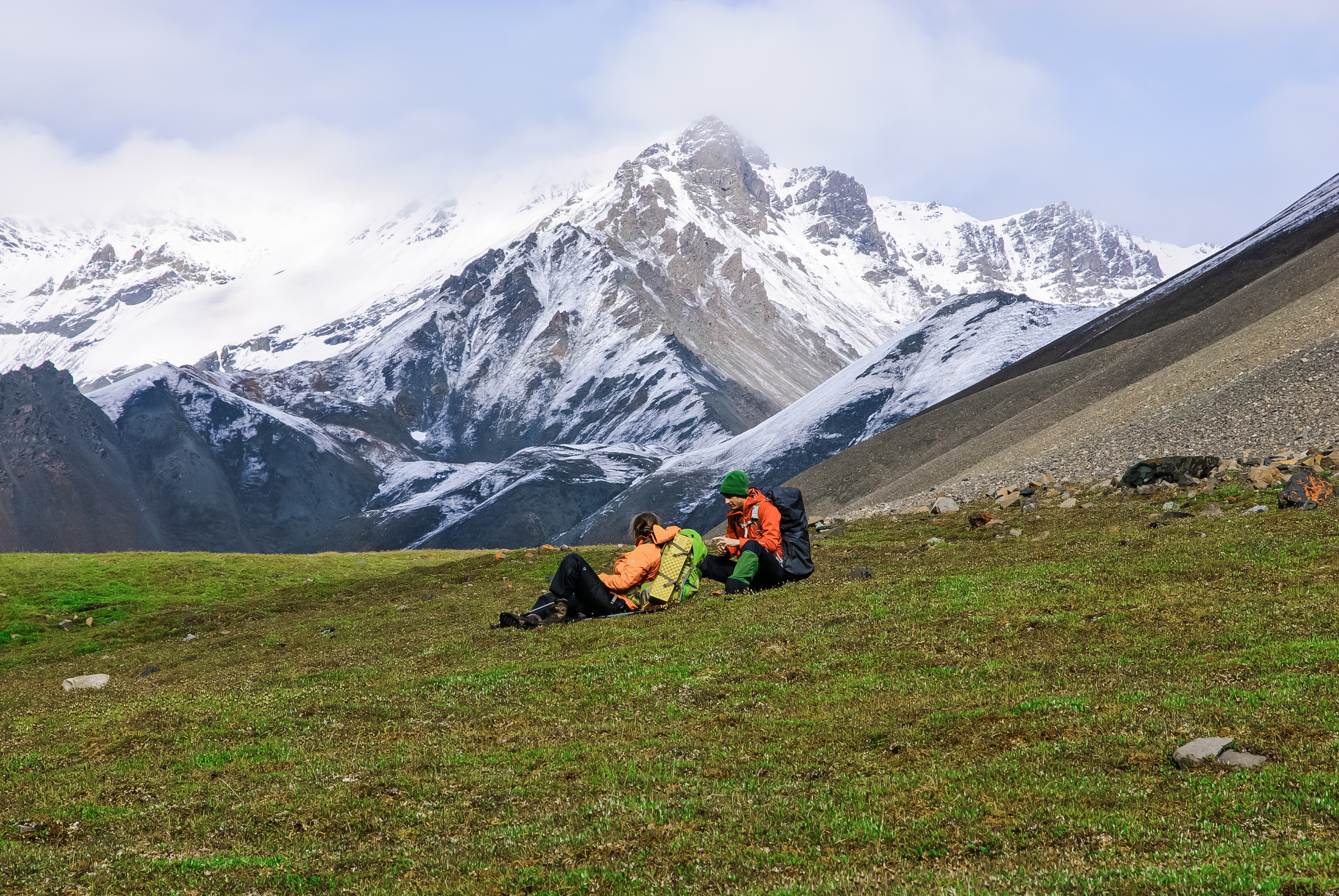 Two backpackers sitting in an alpine meadow with snowy mountains in the background
