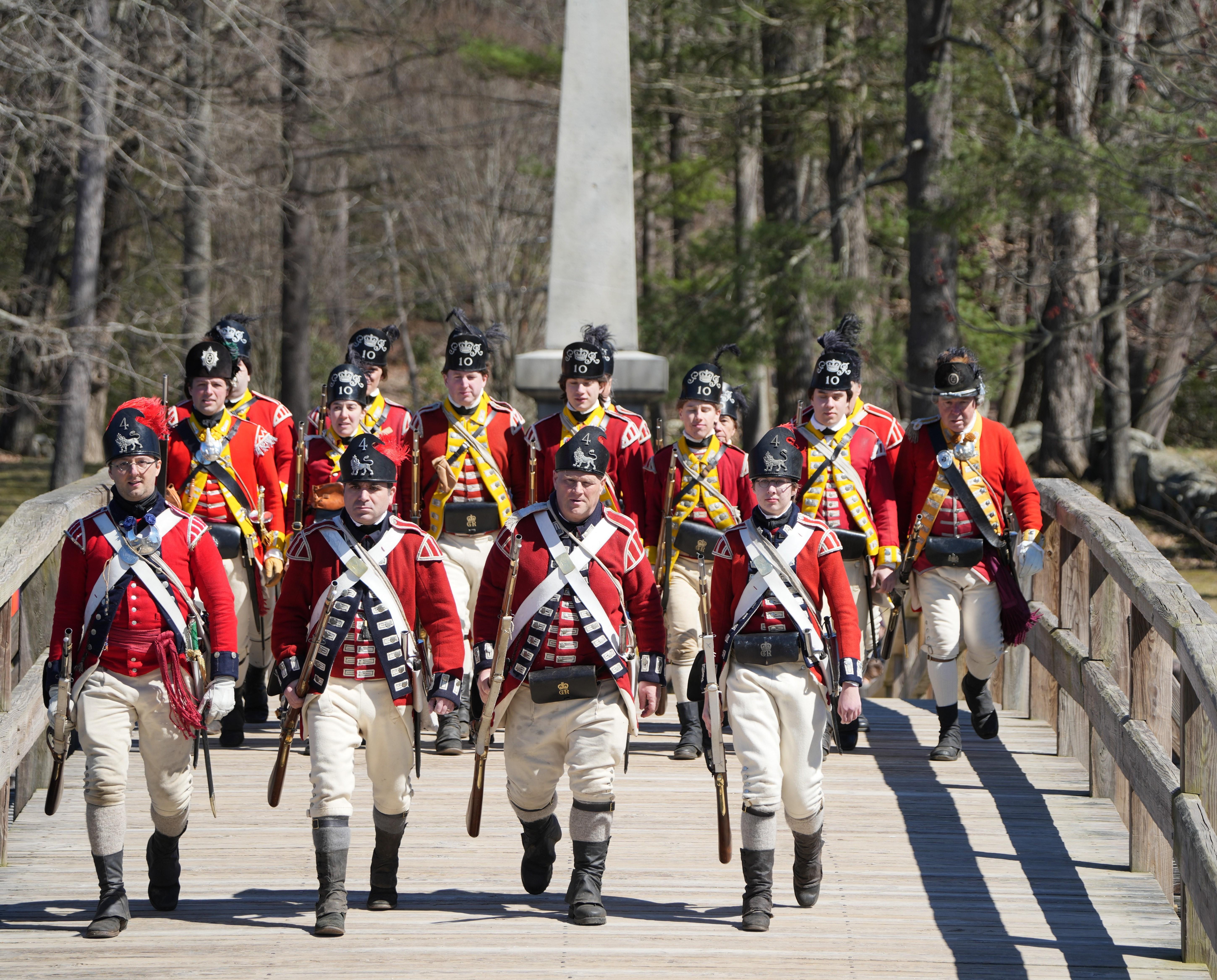 British soldiers marching together over the a wooden bridge. A stone monument towers behind.