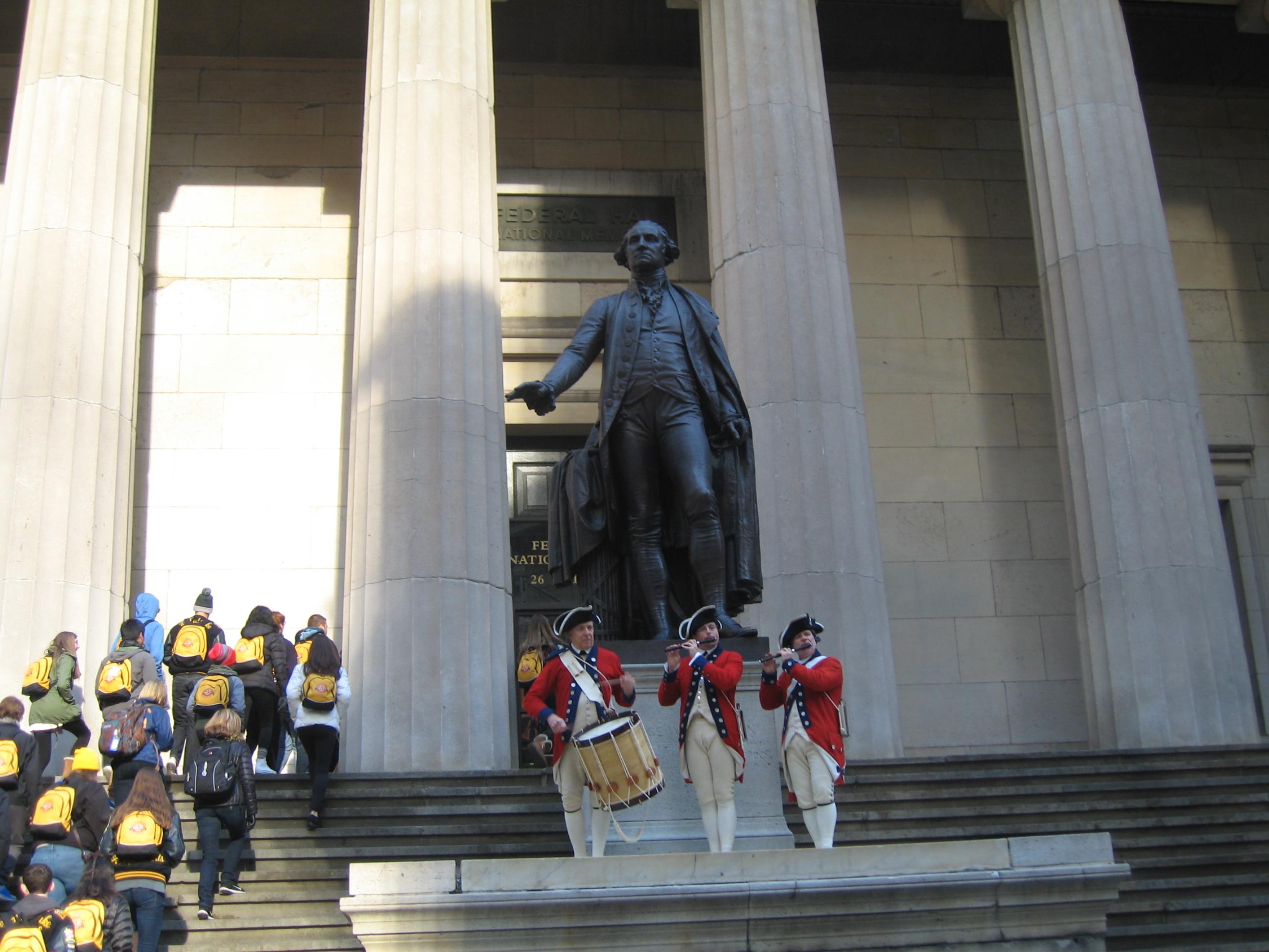Three people in reenactment clothing hold instruments in front of a stone building and giant statue.