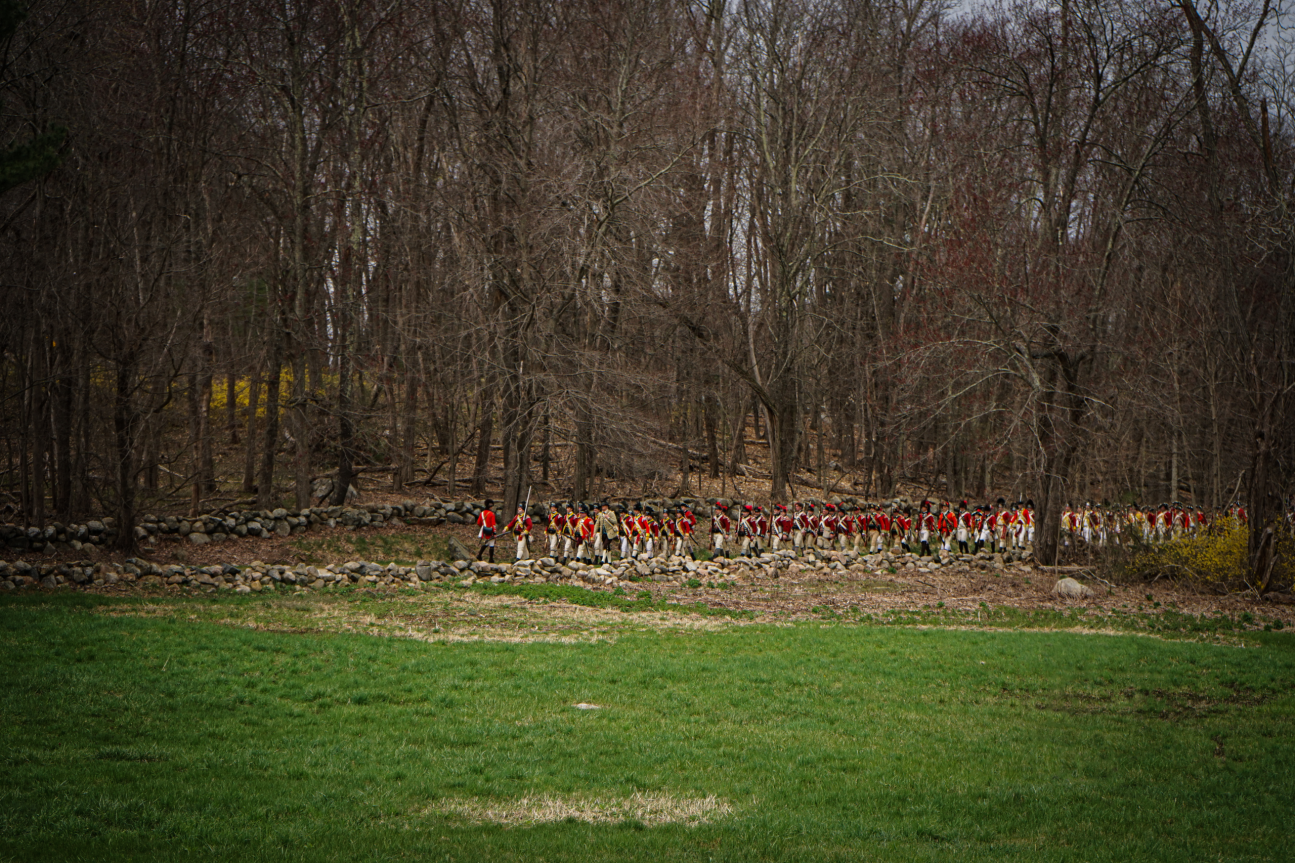 An open field with trees and stone wall. British soldiers in red coats march in the distance.